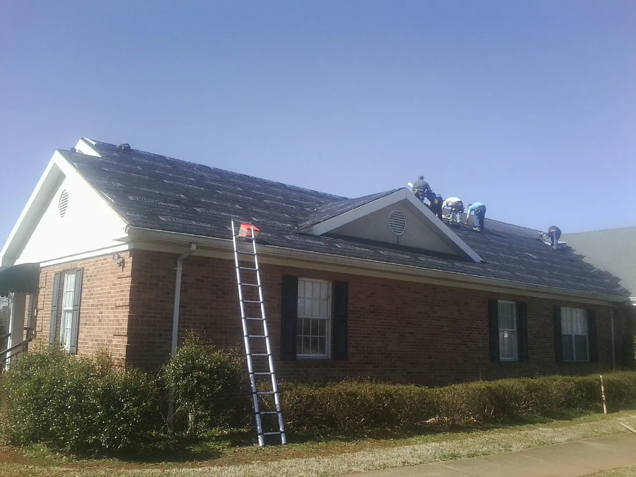Crew tearing shingles off a brick ranch roof with a ladder set — Metal Roof Repair work by our crew in Chelmsford