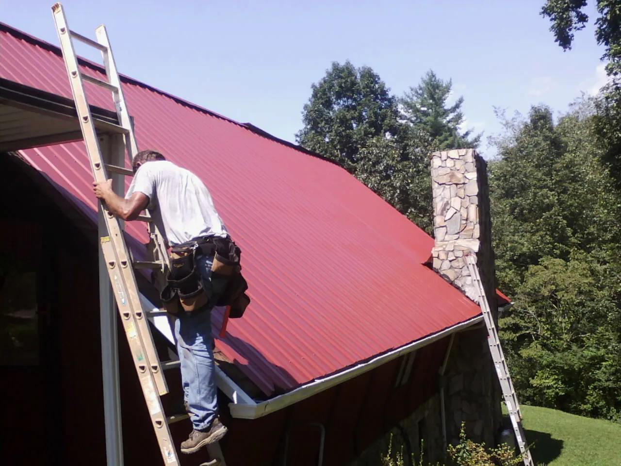 Roofer on a ladder beside a red metal roof and stone chimney — Soffit & Fascia Repair by Roofing Brothers in Chelmsford